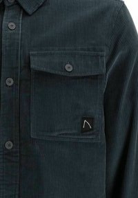 Dark green corduroy shirt with vertical ribbing, featuring button closure and a chest pocket. Black label with a white logo on pocket.