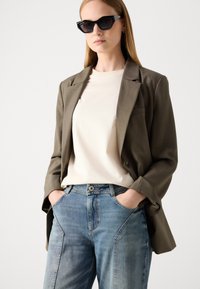 Ελιά πράσινο σακάκι πάνω από ένα κρεμ μπλουζάκι, συνδυασμένο με ανοιχτό μπλε τζιν. Το σακάκι διαθέτει ένα κουμπί και τσέπη στο στήθος. Μαύρα γυαλιά ηλίου.