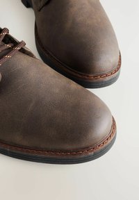Brązowe skórzane buty z gładką, teksturowaną cholewką, okrągłym noskiem i brązowym przeszyciem wzdłuż podeszwy. Gumowa podeszwa zapewniająca przyczepność i trwałość.