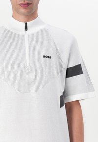 Fehér, rövid ujjú kötött polo, cipzáras gallérral, texturált mintával, fekete részletekkel és "BOSS" logóval a mellkasán.