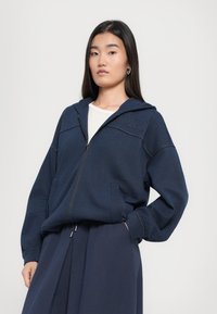 Felpa con zip di colore blu navy realizzata in tessuto morbido, dotata di cappuccio, tasche a canguro e dettagli di cucitura a contrasto. Indossata sopra una maglietta leggera.