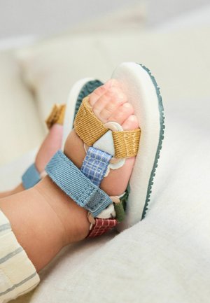 Babyvoeten met kleurrijke stoffen riemsandalen, rustend op een zacht wit oppervlak.