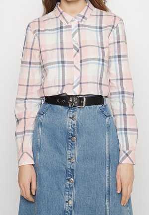 Femme portant une chemise à carreaux rose et bleue boutonnée, rentrée dans une jupe en denim bleu taille haute avec une ceinture noire.
