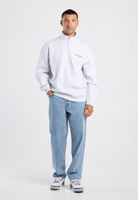 Felpa grigia chiara con zip e polsini a costine, con ricamo "New York". Abbinata a jeans leggeri blu chiaro e scarpe sportive.