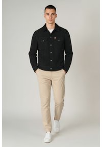 Schwarze Wildlederjacke mit zwei Brusttaschen und vorderen Knöpfen, getragen über einem weißen Hemd, kombiniert mit beiger Hose und weißen Sneakern.