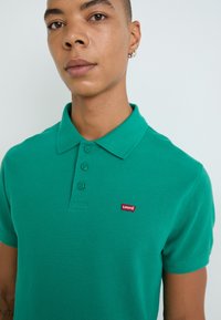 Zelená polo košeľa vyrobená z textúrovaného bavlneného materiálu, s klasickým golierom, trojmiestnym zapínaním na gombíky a červeným logom Levi's na ľavej hrudi.