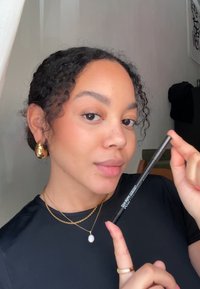Schwarzer Eyelinerstift, gehalten von einer Frau. Sie trägt ein schwarzes Shirt, geschichtete goldene Halsketten und große goldene Creolen. Natürliche Schminke.