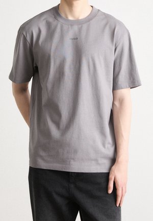 Osoba nosząca gładki szary T-shirt z krótkim rękawem z małym logo "HUGO" na klatce piersiowej oraz czarne spodnie, stojąca z jedną ręką za plecami.