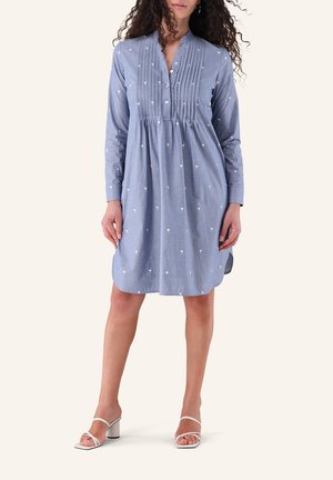 Frau trägt ein locker sitzendes hellblaues Kleid mit langen Ärmeln, kleinen weißen Blumenmustern, Knöpfen vorne und weißen Sandaletten mit Absatz.