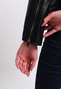 Black leather jacket with a silver zipper detail on the sleeve, showcasing a smooth texture and fitted design. Hand resting on the arm.