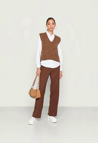 Colete castanho tricotado sobre uma camisa branca, combinado com calças largas castanhas e sapatilhas brancas. Inclui uma pequena bolsa castanha com alça de corrente.