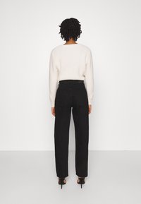 Pantalones negros ajustados con cintura alta y pernera recta, combinados con un suéter de punto color crema, vistos desde atrás. Textura suave y diseño minimalista.