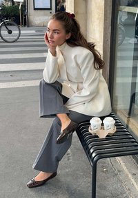 Une personne assise dans un blazer beige et un pantalon gris, portant des mocassins en cuir brun brillant, reposant une main sur son visage ; deux tasses de café sur un banc noir.
