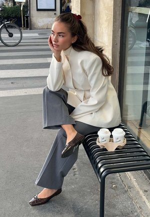 En sittande person i en krämfärgad kavaj och grå byxor, som bär glänsande bruna loafers, med en hand vilande mot ansiktet; två kaffekoppar på en svart bänk.