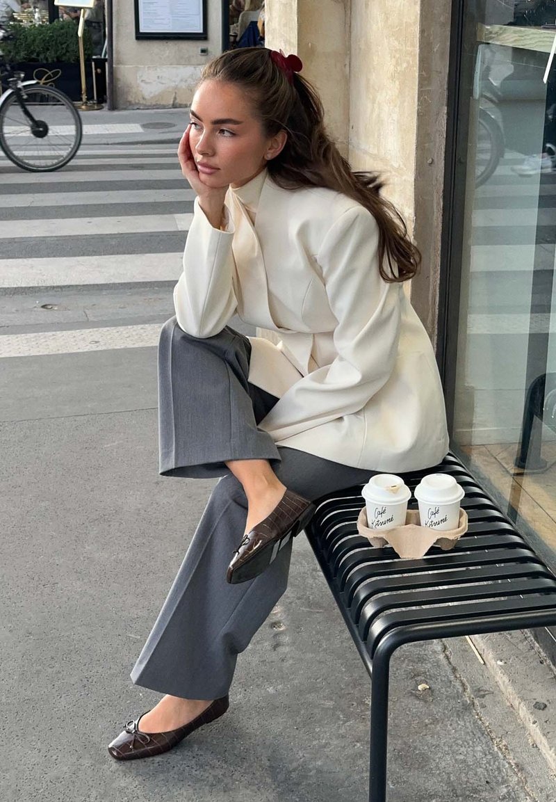 Une personne assise dans un blazer beige et un pantalon gris, portant des mocassins en cuir brun brillant, reposant une main sur son visage ; deux tasses de café sur un banc noir.