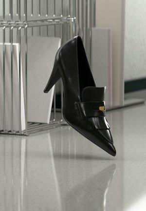 Chaussure à talon haut en cuir noir avec bout pointu et franges décoratives flottant au-dessus d'un sol brillant près d'une étagère métallique avec des documents.