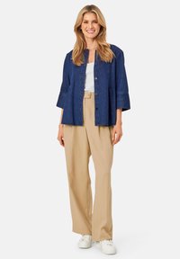 Donkerblauwe denim jacket met een knoopsluiting en driekwart mouwen, gecombineerd met beige wijde broek en witte sneakers.