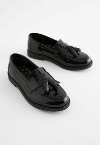 Mocasines de piel sintética negra con suela plana, que cuentan con un detalle de borlas y costuras detalladas a lo largo del empeine y los laterales.