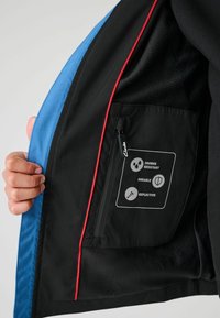 Wasserabweisende Jacke, schwarz mit blauen und roten Akzenten. Verfügt über eine Reißverschlusstasche mit Symbolen, die Eigenschaften anzeigen: duschbeständig, langlebig, reflektierend.