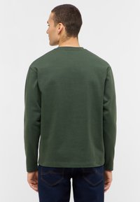 Tamsiai žalia medvilninė ilgomis rankovėmis marškinėliai su apvaliu kaklu, reguliaraus kirpimo, lygia tekstūra ir minimalistiniu dizainu.