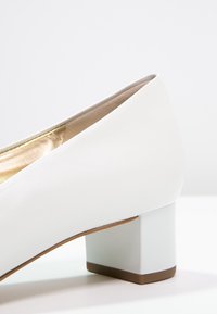 Chaussure blanche à petit talon bloc, texture lisse et doublure intérieure dorée sur fond blanc.