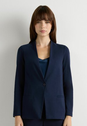 Donna con capelli castani che indossa un blazer blu navy sopra una maglia abbinata, in piedi davanti a una parete liscia di colore grigio chiaro.