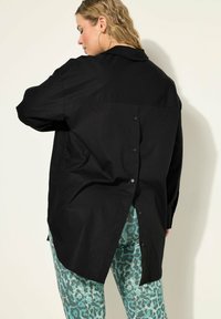 Sort button-up skjorte med løs pasform, der har en rygsplit detalje, glat tekstur og lange ærmer, parret med mønstrede tealfarvede leggings.