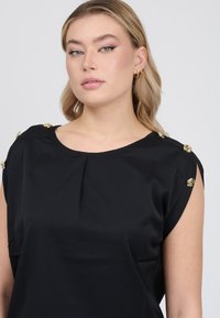 Top nero a maniche corte con dettagli in hardware dorato sulle spalle, tessuto liscio, scollatura rotonda e leggero plissè anteriore.