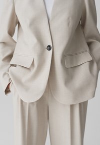 Beige blazer met gestructureerde pasvorm, enkele zwarte knoopsluiting en twee voorzakken. Geweven stof met subtiele patroonDetails.