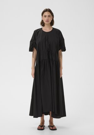 Femme aux cheveux courts et bruns porte une robe midi noire ample avec des manches froncées et des sandales noires, debout sur un fond gris uni.