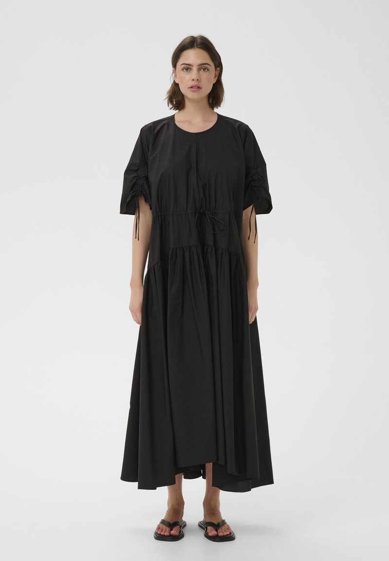 Femme aux cheveux courts et bruns porte une robe midi noire ample avec des manches froncées et des sandales noires, debout sur un fond gris uni.