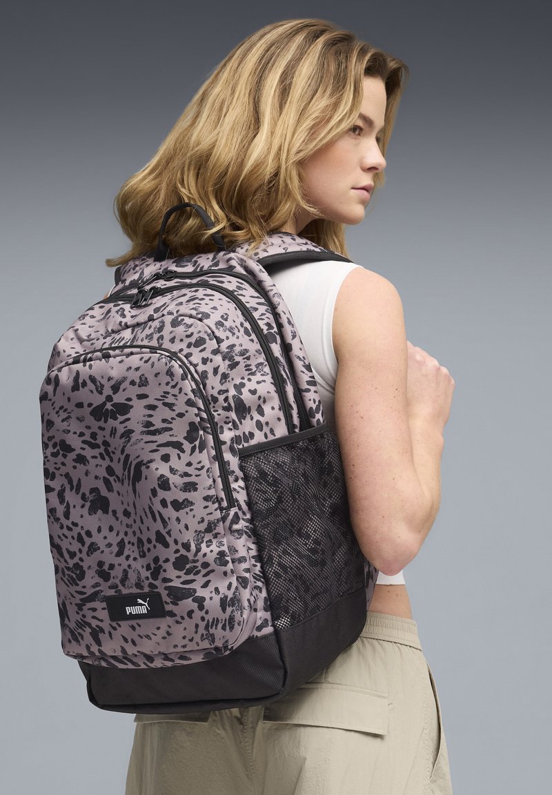 Ung kvinde med skulderlangt blondt hår iført en lilla og sort mønstret Puma-rygsæk, hvid ærmeløs top og beige bukser.