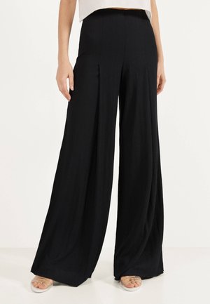 Mujer que lleva pantalones negros de talle alto y pierna ancha, de pie con las manos relajadas a los lados, combinados con un top corto blanco y sandalias.