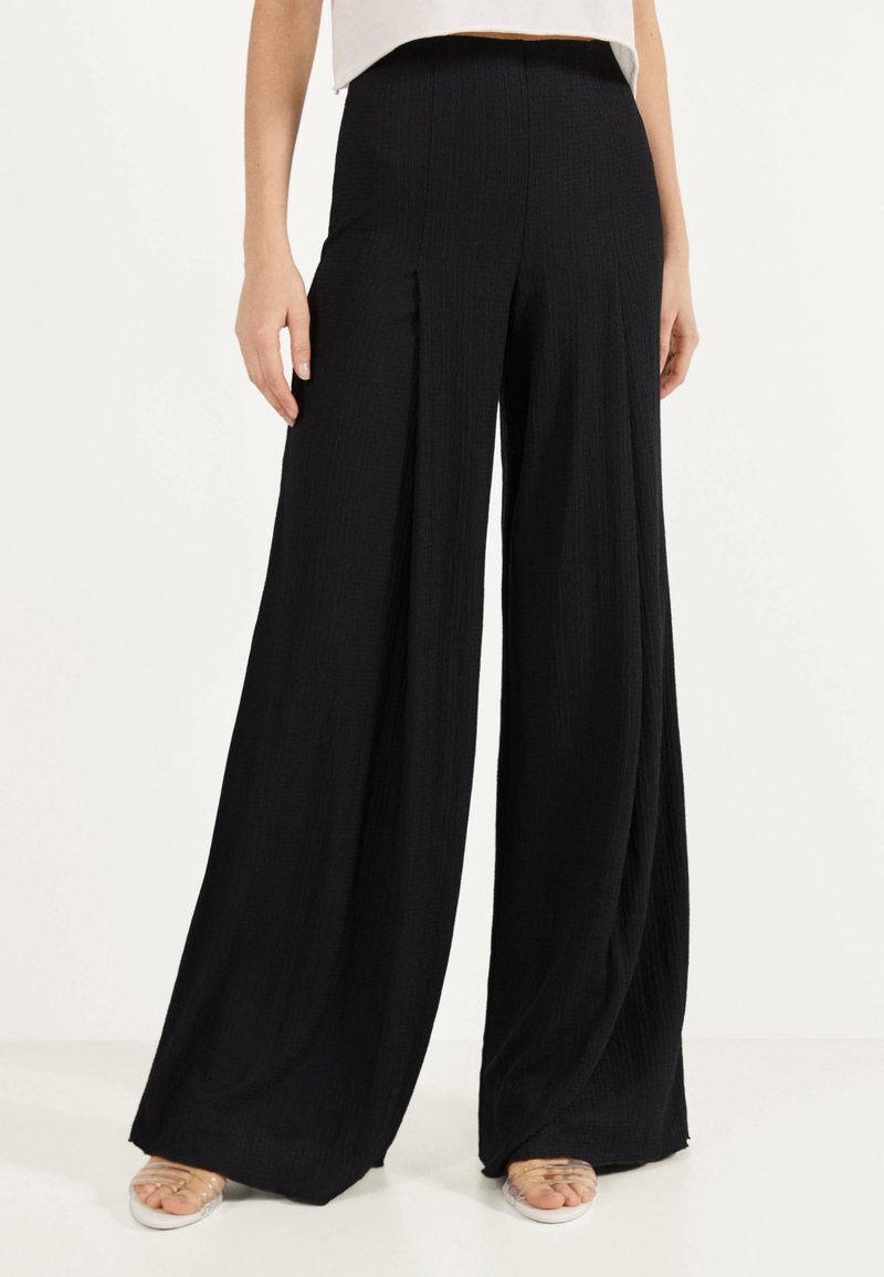 Femme portant un pantalon noir taille haute à jambes larges, debout, les mains détendues le long du corps, associé à un top court blanc et des sandales.