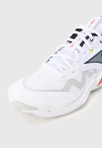 Λευκό αθλητικό παπούτσι με άνω μέρος από πλέγμα και συνθετικό υλικό, χαρακτηριστικό κυματιστό σχέδιο, μαύρες και γκρι λεπτομέρειες και κόκκινα στοιχεία στη σόλα.