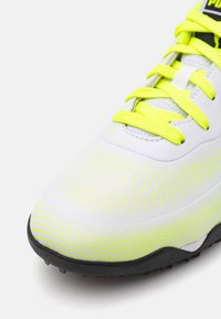 Bota de fútbol blanca con superficie texturada, detalles amarillos y suela de goma negra con tacos de tracción. Los cordones son de un amarillo brillante.