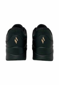 Ein Paar schwarze Sneakers mit strukturierten Sohlen, von hinten betrachtet, mit einem goldenen stilisierten "S"-Logo an jeder Ferse.