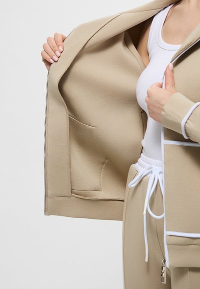 Giacca beige con zip in tessuto liscio, dotata di tasche laterali e dettagli bianchi, abbinata a una canotta bianca e pantaloni beige coordinati.