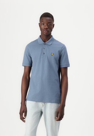 Cămașă polo din bumbac albastru, cu guler și trei nasturi, având un logo mic brodat galben pe piept, combinată cu blugi deschiși din denim.