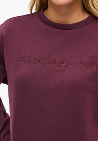 Donna con una felpa color bordeaux con la parola "MINIMALISMO" ricamata sul petto con filo coordinato.