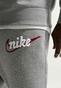 Prim-plan cu o persoană care poartă pantaloni de trening gri și hanorac cu logo-ul Nike brodat alb și roșu pe coapsă.