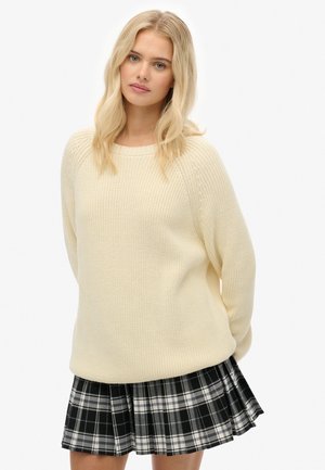 Blonde woman wearing a cream knit sweater and black-white plaid pleated skirt, standing with hands behind back against white background.