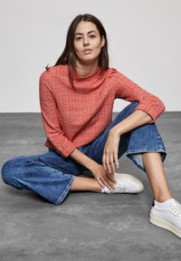Maglione rosso con texture e collo alto, abbinato a jeans blu a gamba larga e scarpe da ginnastica bianche. Tessuti morbidi e vestibilità rilassata.