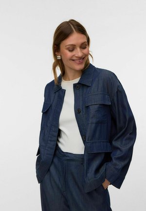 Naine naeratab ja vaatab alla, kandes tumesinist nööpidega jopet esitaskutega valge topi ja sobivate pükstega.