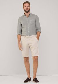 Lichtgrijs shirt met lange mouwen en een zak, gecombineerd met beige shorts en donkerbruine instappers. Eenvoudige, casual outfit tegen een neutrale achtergrond.