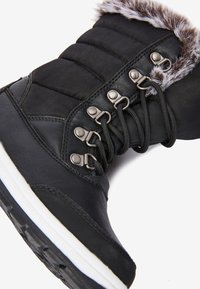 Stivale invernale nero che presenta un mix di pelle liscia e nylon texture. Ha occhielli in metallo, lacci e un colletto in pelliccia sintetica. Suola bianca.