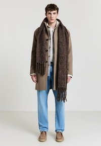 Cappotto di lana marrone con bottoni, indossato sopra una camicia chiara. Una sciarpa frangiata marrone abbinata, jeans azzurri chiari e scarpe color beige completano l'outfit.