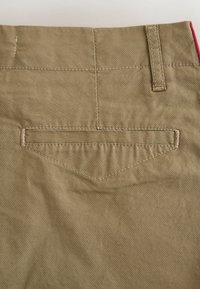 Poche arrière d’un pantalon en tissu beige avec des coutures détaillées et une seule passant de ceinture au-dessus de la poche, bord de doublure rouge visible en haut à droite.