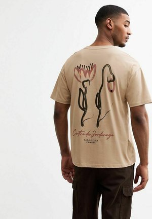 Beżowy t-shirt z bawełny z floralnym nadrukiem w czerni i różu z tyłu, z napisem "Centre de Jardins" na dole.