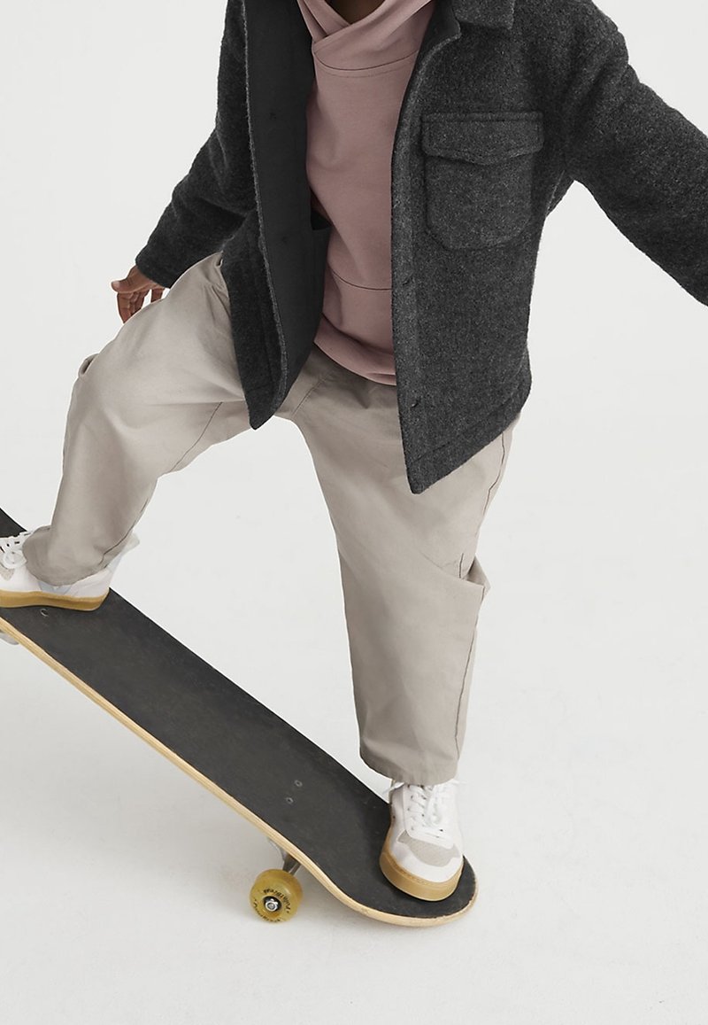 Personne portant un pantalon beige, des baskets blanches, un sweat à capuche rose et une veste gris foncé, en équilibre sur un skateboard avec un pied sur la planche.